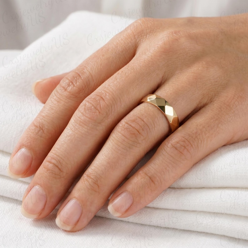 May include: A gold ring with a geometric, faceted design. The ring is a warm gold color and has a series of flat, angled surfaces that catch the light. The ring is on a finger, resting on a white cloth.
