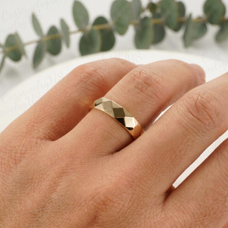 May include: A gold-colored ring with a geometric, faceted design. The ring is worn on a finger, showcasing its polished surface and the play of light across its angles. The ring's design is modern and minimalist, suitable for everyday wear.