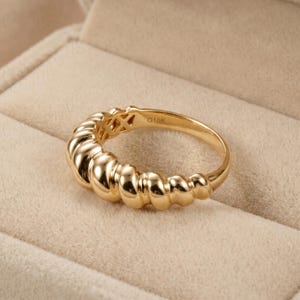 May include: A gold ring with a textured, ridged design. The ring has a smooth, polished finish and features the inscription "G18K" on the inner band. The ring is set against a neutral background.