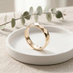 May include: A gold ring with a geometric faceted design. The ring is set on a white ceramic plate, with a sprig of green leaves in the background. The ring is marked "14K".