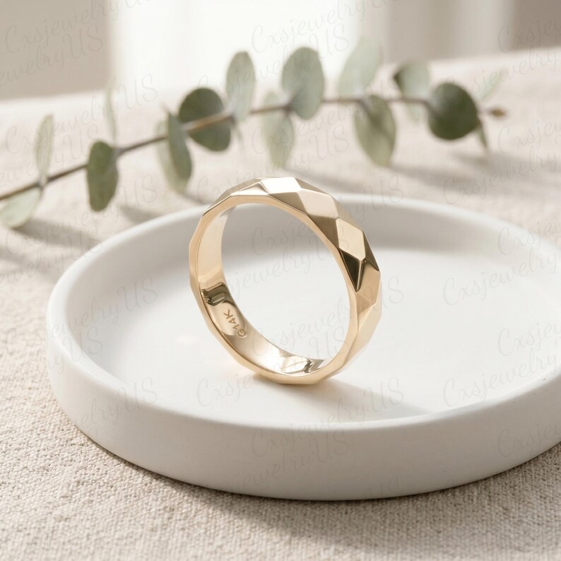 May include: A gold ring with a geometric faceted design. The ring is set on a white ceramic plate, with a sprig of green leaves in the background. The ring is marked "14K".