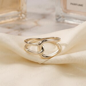 May include: A gold-colored ring with a unique, intertwined design. The ring features a smooth, polished surface and a double-banded structure, creating an eye-catching, modern look. The ring is displayed on a cream-colored fabric.