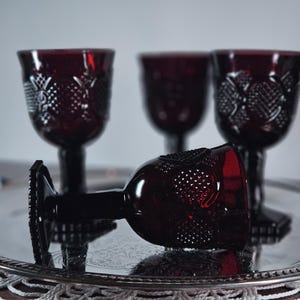 May include: Four ruby red vintage wine glasses with ornate detailing, one lying on its side on a silver serving tray. The glasses have a textured pattern and a dark stem. The background is a neutral color.