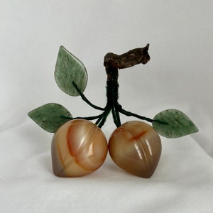 May include: A carved agate fruit sculpture with two peach-colored fruits and green leaves. The sculpture is made of natural stone and has a rustic, handmade look.