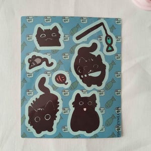 Könnte beinhalten: Ein rechteckiger Aufkleberbogen mit hellblauem Hintergrund und einem sich wiederholenden Muster aus kleinen Fischsymbolen. Der Bogen enthält mehrere dunkelbraune Katzenaufkleber, darunter Katzen in verschiedenen Posen, eine Spielzeugmaus und ein Fischspielzeug.