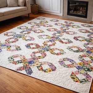 May include: A large, floral-patterned quilt with a cream-colored background. The quilt features a repeating design of interlocking shapes filled with colorful flowers, including roses and sunflowers. The quilt is placed on a wooden floor.