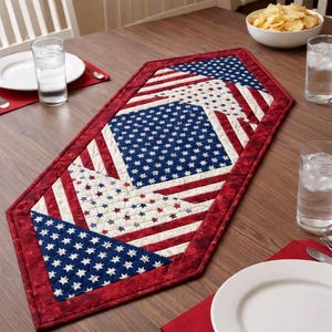 May include: A patriotic table runner with a red, white, and blue design. The runner features a patchwork pattern with stars and stripes, reminiscent of the American flag. The edges are trimmed with a dark red fabric.