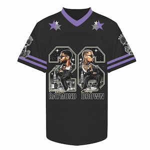 May include: A black jersey with purple V-neck trim and sleeve stripes. The front features two large portraits of Usher Raymond and Chris Brown over a city skyline, with the names printed below. Two star icons appear near the neckline.