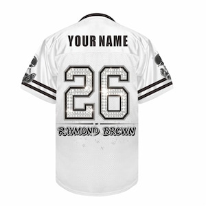 May include: A white sports jersey featuring the text YOUR NAME at the top, the number 26 with a diamond-like pattern in the center, and the name RAYMOND BROWN printed below. The sleeves have black stripes and a graphic of a trophy on each side.
