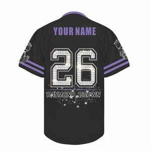 May include: A black baseball jersey with purple trim on the collar and sleeves. The back features the text YOUR NAME, the number 26 in a silver diamond-like pattern, and the name RAYMOND BROWN with a graphic of white stars and sparkles.