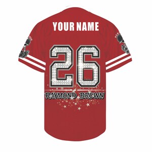 May include: A red baseball jersey featuring the text YOUR NAME in white, the number 26 in a silver diamond-like pattern, and the text RAYMOND BROWN in black. The sleeves have white stripes, and small graphic illustrations appear on each shoulder.
