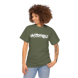 May include: An olive green t-shirt featuring the white word "UNSTOPPABLE". The text has a stylized design. The t-shirt has short sleeves and a crew neck. The model is wearing blue jeans.