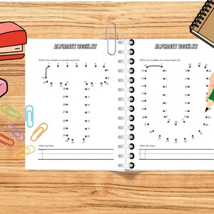 May include: An open spiral-bound alphabet booklet with the text "ALPHABET BOOKLET" is shown. A hand is holding a green pencil, connecting dots to form the letter "U". School supplies like a stapler, eraser, and paperclips are also visible.
