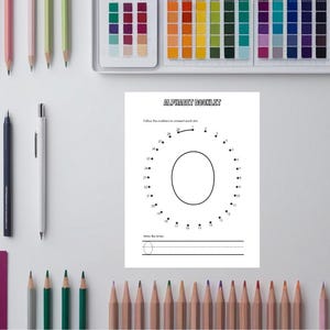 May include: A white worksheet with the text "ALPHABET BOOKLET" and a dot-to-dot letter "O" is surrounded by colored pencils, pens, and a tablet. The worksheet has lines for writing the letter.