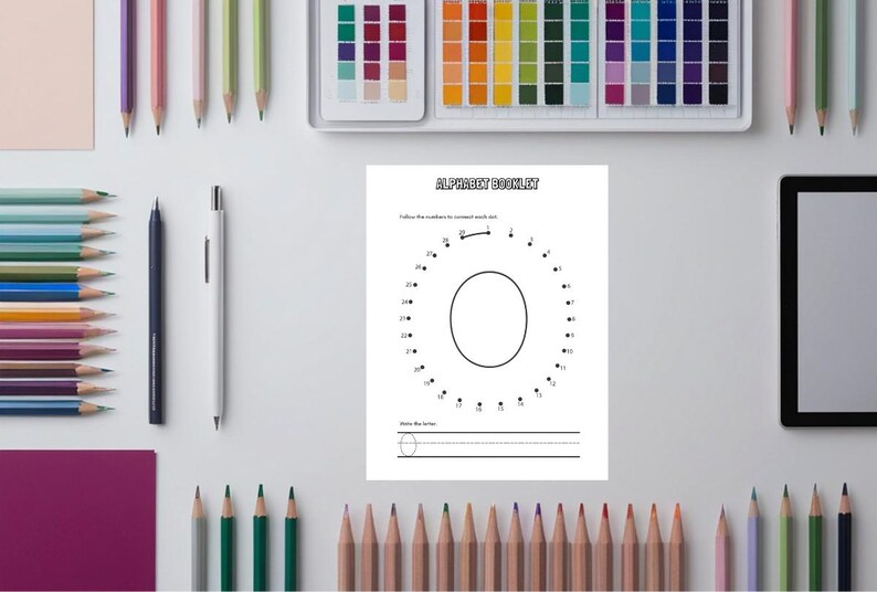 May include: A white worksheet with the text "ALPHABET BOOKLET" and a dot-to-dot letter "O" is surrounded by colored pencils, pens, and a tablet. The worksheet has lines for writing the letter.