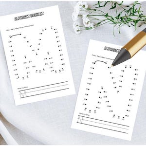 May include: Two white alphabet booklet pages with dot-to-dot letter M exercises. The text "ALPHABET BOOKLET" is at the top. A black and gold pen rests on the pages. White flowers are in the background.