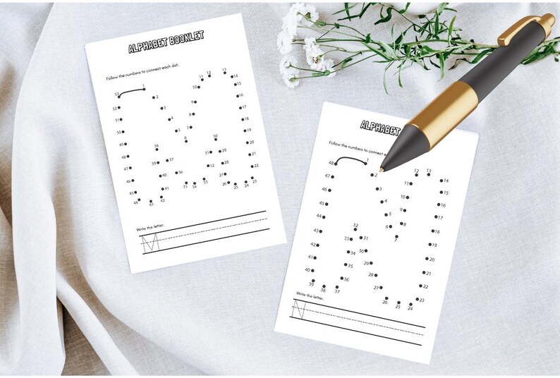 May include: Two white alphabet booklet pages with dot-to-dot letter M exercises. The text "ALPHABET BOOKLET" is at the top. A black and gold pen rests on the pages. White flowers are in the background.