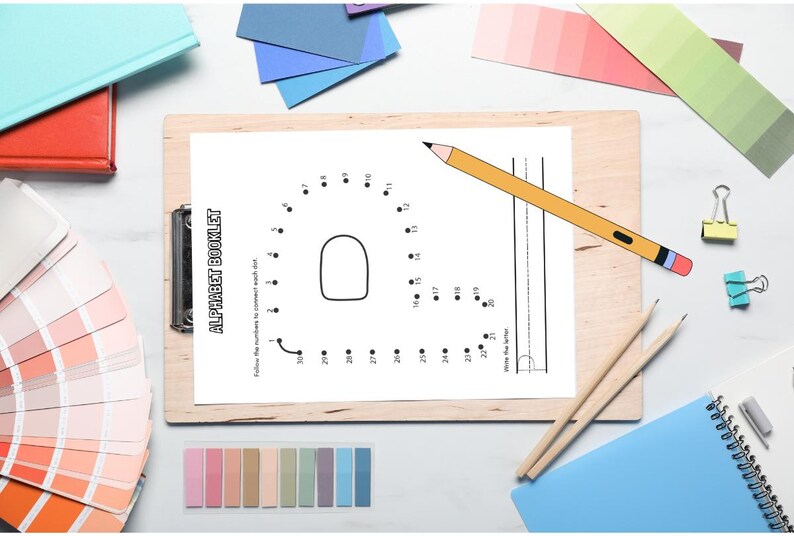 May include: A wooden clipboard holds a white page from an alphabet booklet with the letter "A" as a connect-the-dots activity. A yellow pencil rests on the page. Other stationery items, including colored paper swatches, are scattered around.