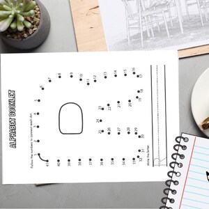 May include: A black and white alphabet booklet with the text "ALPHABET BOOKLET" and a dot-to-dot letter. A cup of coffee, a notepad with a pencil, and a succulent plant are also visible.
