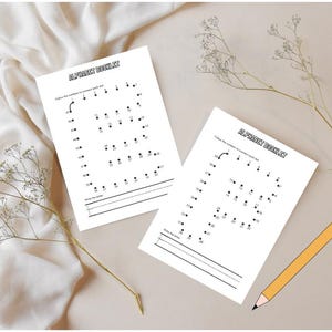 May include: Two white worksheets with the text "ALPHABET BOOKLET" and dot-to-dot letters are displayed on a beige surface. A yellow pencil and a green pencil holder with pencils are also present. The background is a white fabric.