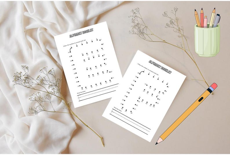 May include: Two white worksheets with the text "ALPHABET BOOKLET" and dot-to-dot letters are displayed on a beige surface. A yellow pencil and a green pencil holder with pencils are also present. The background is a white fabric.