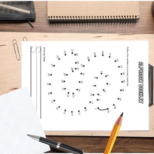 May include: A white paper with the text "ALPHABET BOOKLET" and a dot-to-dot puzzle. The image includes a pencil, pens, a notepad, and a cup of coffee on a wooden surface. The dot-to-dot puzzle is numbered 1-49.