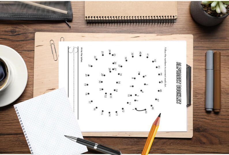 May include: A white paper with the text "ALPHABET BOOKLET" and a dot-to-dot puzzle. The image includes a pencil, pens, a notepad, and a cup of coffee on a wooden surface. The dot-to-dot puzzle is numbered 1-49.