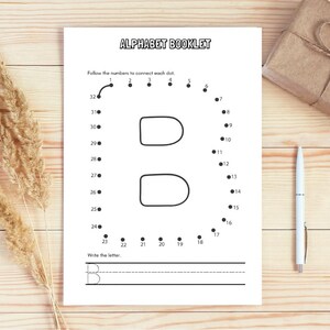 May include: A white alphabet booklet with the letter "B" as a connect-the-dots activity. The booklet is on a wooden surface, with a pen and a wrapped gift. Dried wheat stalks are on the left.