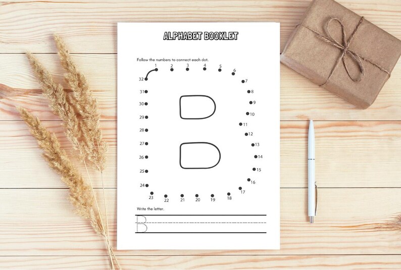 May include: A white alphabet booklet with the letter "B" as a connect-the-dots activity. The booklet is on a wooden surface, with a pen and a wrapped gift. Dried wheat stalks are on the left.