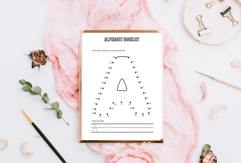 May include: A framed alphabet booklet with the letter "A" as a connect-the-dots activity. The booklet is on a white surface with pink fabric, a dried rose, and a paintbrush. The text "ALPHABET BOOKLET" is at the top.