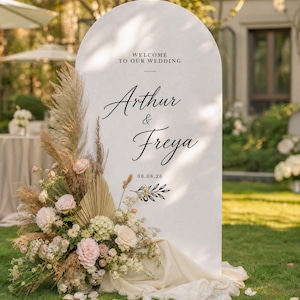May include: A white arched wedding welcome sign featuring the black text "WELCOME TO OUR WEDDING", "Arthur & Freya", and "08.08.26". The sign is decorated with a large arrangement of pampas grass, dried palm leaves, and light pink and white flowers.
