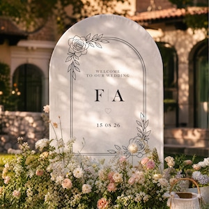 Può includere: Un cartello di benvenuto per matrimoni bianco ad arco con disegni a linee nere di rose e foglie. Il testo recita Welcome to our wedding, le iniziali F A e la data 15 08 26. Il cartello è circondato da una fitta composizione di fiori bianchi e rosa.