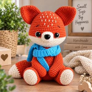 May include: A crocheted fox stuffed animal with bright orange fur, a white muzzle, and a white-tipped tail. The fox features large black safety eyes, embroidered white forehead details, and wears a textured, vibrant blue crocheted scarf.