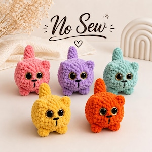 May include: Five small, round, crocheted plush cat figures in pink, purple, light blue, yellow, and orange. Each figure features large black safety eyes, a stitched black mouth, and a small upright tail. The text No Sew is visible above.