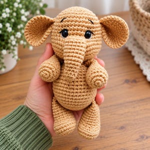 Könnte beinhalten: Ein hellbraunes Häkel-Elefant-Spielzeug mit rundem Kopf, großen Ohren, einem zentralen Rüssel und zwei schwarzen Kunststoffaugen mit gestickten Augenbrauen. Das Stofftier hat eine strukturierte Garnoberfläche und einen kompakten Körper.