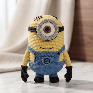 Puede incluir: Una figura amigurumi de ganchillo de un personaje amarillo con un solo ojo grande, que lleva un peto azul y guantes negros. La figura presenta un marco de ojo gris y blanco, un iris marrón, una pupila negra y una pequeña sonrisa negra.
