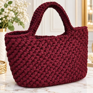 Può includere: Una borsa a mano color bordeaux intenso realizzata con una spessa trama di corda intrecciata. La borsa presenta un design strutturato con apertura superiore e due manici integrati, caratterizzato da un motivo a nodi costante e ripetitivo.