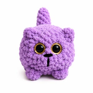 May include: A round, purple crochet plush toy shaped like a cat. The figure features a textured, yarn-based surface, two large black eyes with gold-colored rims, a small black stitched nose and mouth, and a short, upright tail on top.