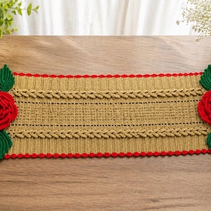 Puede incluir: Un camino de mesa rectangular tejido a mano. El camino de mesa es principalmente beige con un borde rojo festoneado. Presenta un diseño trenzado y está adornado con rosas rojas y hojas verdes en cada extremo.