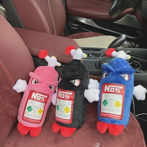 May include: Three plush toys shaped like NOS bottles in pink, black, and blue. Each has a white and red top, white hands, and red feet. The bottles have the "NOS" logo and a label that reads "Nitrous Oxide Systems."