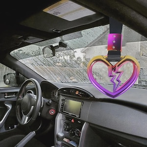 May include: A car interior with a decorative heart-shaped handle. The handle is a gradient of gold, pink, and purple, with a lightning bolt design. The car's dashboard, steering wheel, and windshield are also visible.