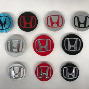 May include: A collection of ten Honda wheel center caps in various colors. The caps feature the Honda logo in silver, red, black, and blue. The caps are round and have a metallic finish.