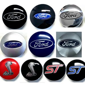 May include: A collection of Ford car emblems in various colors, including black, silver, blue, and red. The emblems feature the Ford logo, a cobra, and ST lettering. The emblems are round and appear to be made of metal.