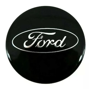 May include: A black, circular Ford emblem with a glossy finish. The word "Ford" is written in a stylized, silver font within a white oval. The emblem is likely for a car.