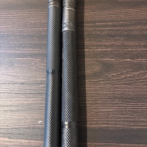May include: Two black metal pen-like objects with a textured, diamond-patterned grip. The objects are cylindrical and have a slightly darker top section. They are positioned vertically against a dark wood grain surface.
