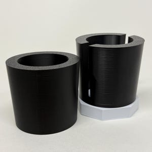 May include: Two black cylindrical objects with a textured surface and a gray foam rim. One is a complete cylinder, the other is a cylinder with a section removed, sitting on a white hexagonal base. The objects are on a white background.