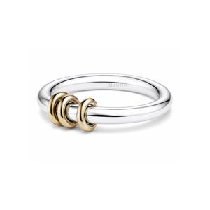 Puede incluir: Un anillo plateado con tres anillos entrelazados de color dorado. El anillo tiene un acabado liso y pulido, y el nombre de la marca "BJORA" está grabado en la banda. El diseño es minimalista y moderno.