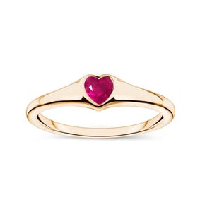 May include: A gold ring with a heart-shaped, bright red gemstone. The ring's band is smooth, polished gold, and the heart-shaped stone is set in a bezel setting. A delicate piece of jewellery.