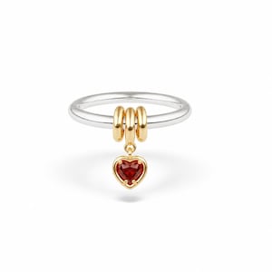 Op de afbeelding: Een zilveren ring met drie goudkleurige ringetjes die aan de band zijn geregen. Een kleine goudkleurige hartvormige bedel met een donkerrode edelsteen in het midden hangt aan de middelste ring, wat een opvallend accent geeft aan de ring.