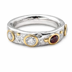 Puede incluir: Un anillo de banda de plata texturizada con una serie de piedras engastadas, incluyendo dos piedras redondas facetadas transparentes y una piedra redonda facetada de color rojo oscuro. La banda tiene un acabado martillado con detalles dorados.